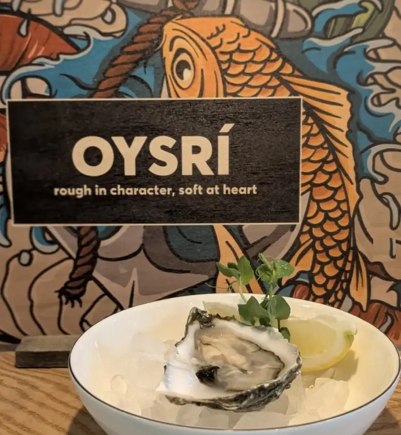 Oysrí oyster on ice with lemon in front of the wooden sign “Oysrí – rough in character, soft at heart”
